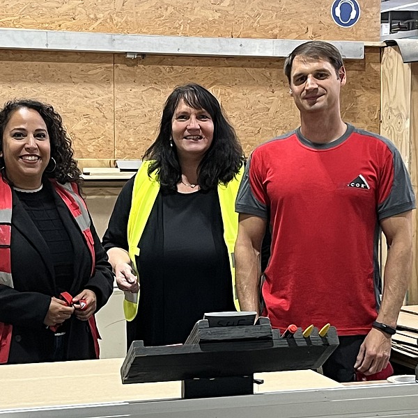 Drei Personen stehen lächelnd in einer Werkhalle vor einer Maschine; zwei tragen Arbeitskleidung, eine trägt eine gelbe Sicherheitsweste