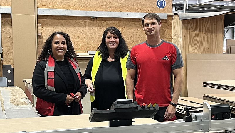 Drei Personen stehen lächelnd in einer Werkhalle vor einer Maschine; zwei tragen Arbeitskleidung, eine trägt eine gelbe Sicherheitsweste