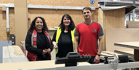 Drei Personen stehen lächelnd in einer Werkhalle vor einer Maschine; zwei tragen Arbeitskleidung, eine trägt eine gelbe Sicherheitsweste