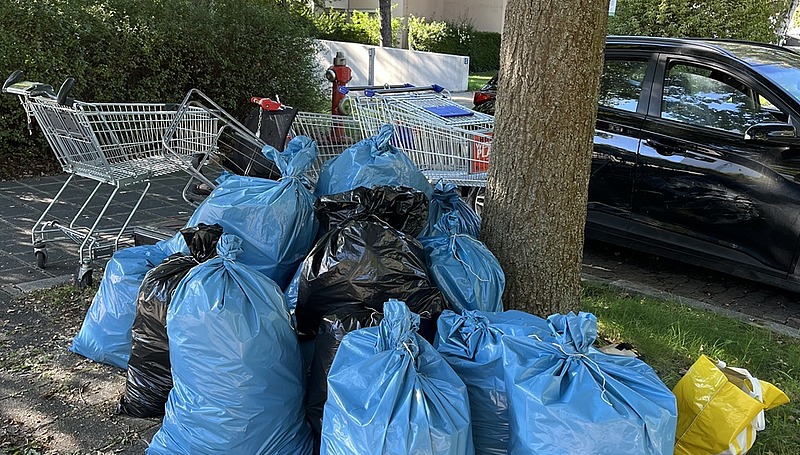 Mehrere blaue Müllsäcke stehen gesammelt unter einem Baum, daneben liegen weitere abgelegte Gegenstände im Freien.